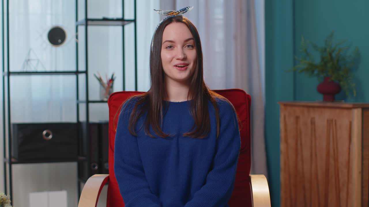 Young woman smiling friendly at camera waving hands gesturing hello hi greeting at home on chair