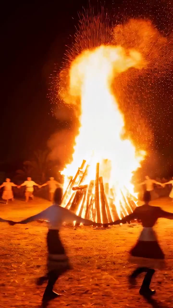 Video vertical: Hoguera chispeante, bailarines con faldas formando un círculo y tomados de la mano en un ritual nocturno.