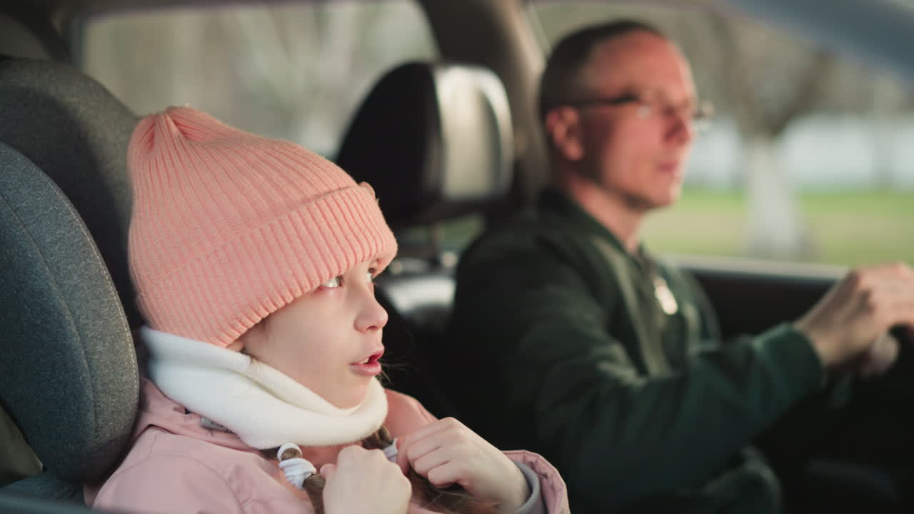 핑크색 모자와 재을 입은 어린 소녀가 차 앞 승객 좌석에 앉아서 손을 움직여 노래하고 있다.