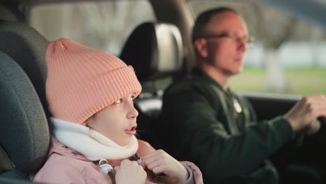 a young girl wearing a pink cap and jacket is sitting in the front passenger seat of a car, singing with her hands in motion. beside her, a man in the driver s seat, wearing glasses and a green jacket