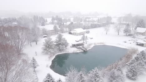 Icy-water-pond-near-small-town-in-United-States-during-heavy-snowfall,-aerial-view