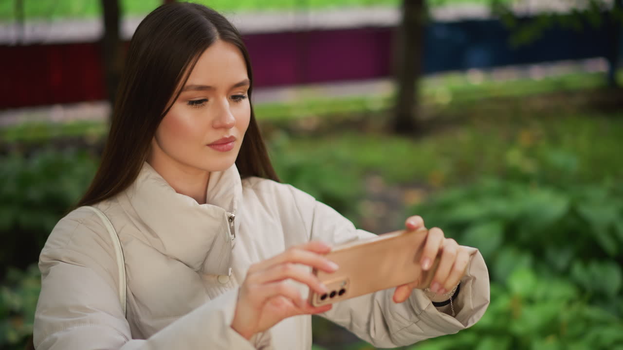 Giovane donna che scatta un selfie all'aperto con cappotto beige e smartphone, modifica l'inquadratura, controlla lo schermo e registra espressioni delicate in un parco diurno con vegetazione verde, influencer rilassata