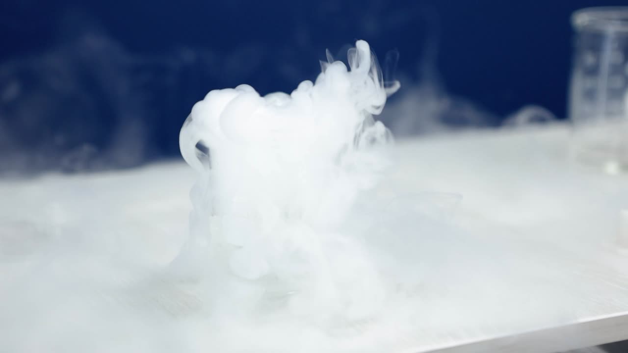 A beaker in a laboratory emits a cascade of white smoke, bubbling and swirling mysteriously against a blue background