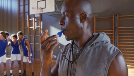 african american male basketball coach using whistle with his team in background