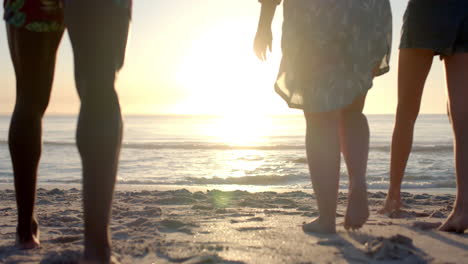 Diverse-four-people-stand-on-a-sandy-beach-at-sunset,-waves-gently-lapping-at-their-feet