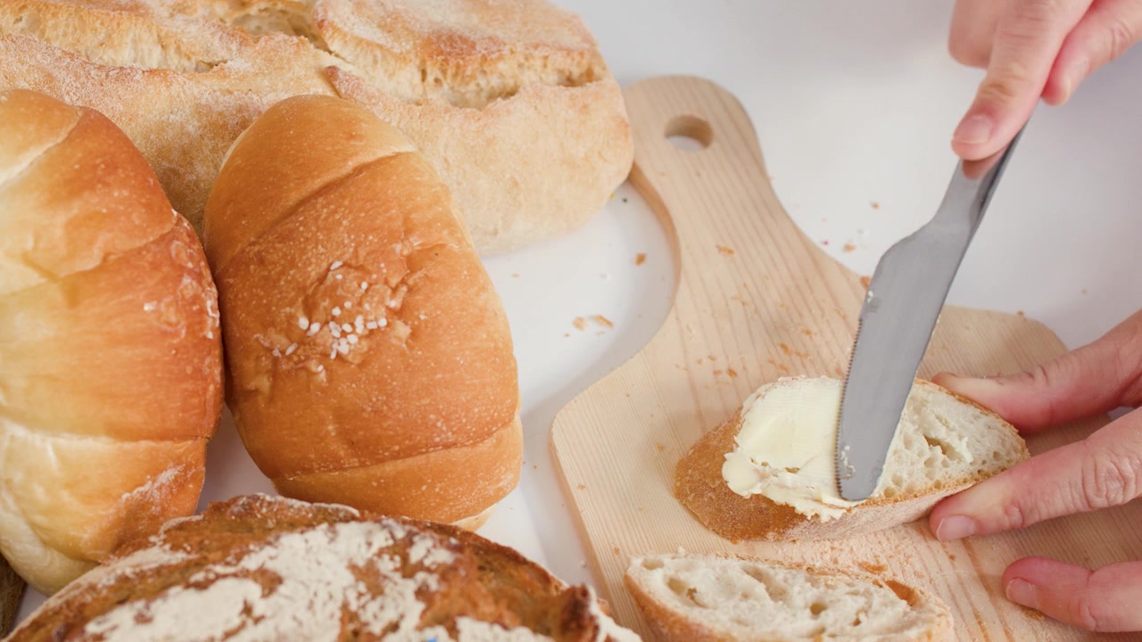 A person spreads creamy butter onto fresh baguette slices atop a wooden cutting board.