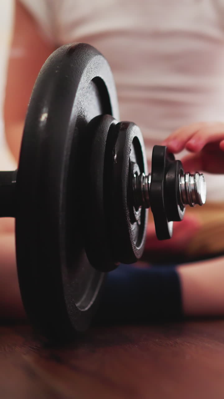 Bored little boy spins gear with hand on big heavy barbell. Toddler sits on floor in gym examining workout equipment closeup slow motion