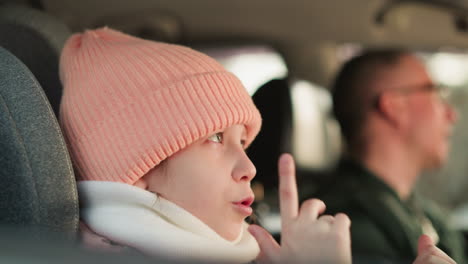 a young girl wearing a pink beanie and jacket is seated in the car, as she gestures with her hands.a blurred man, wearing glasses and a green jacket, is visible in the driver's seat, singing