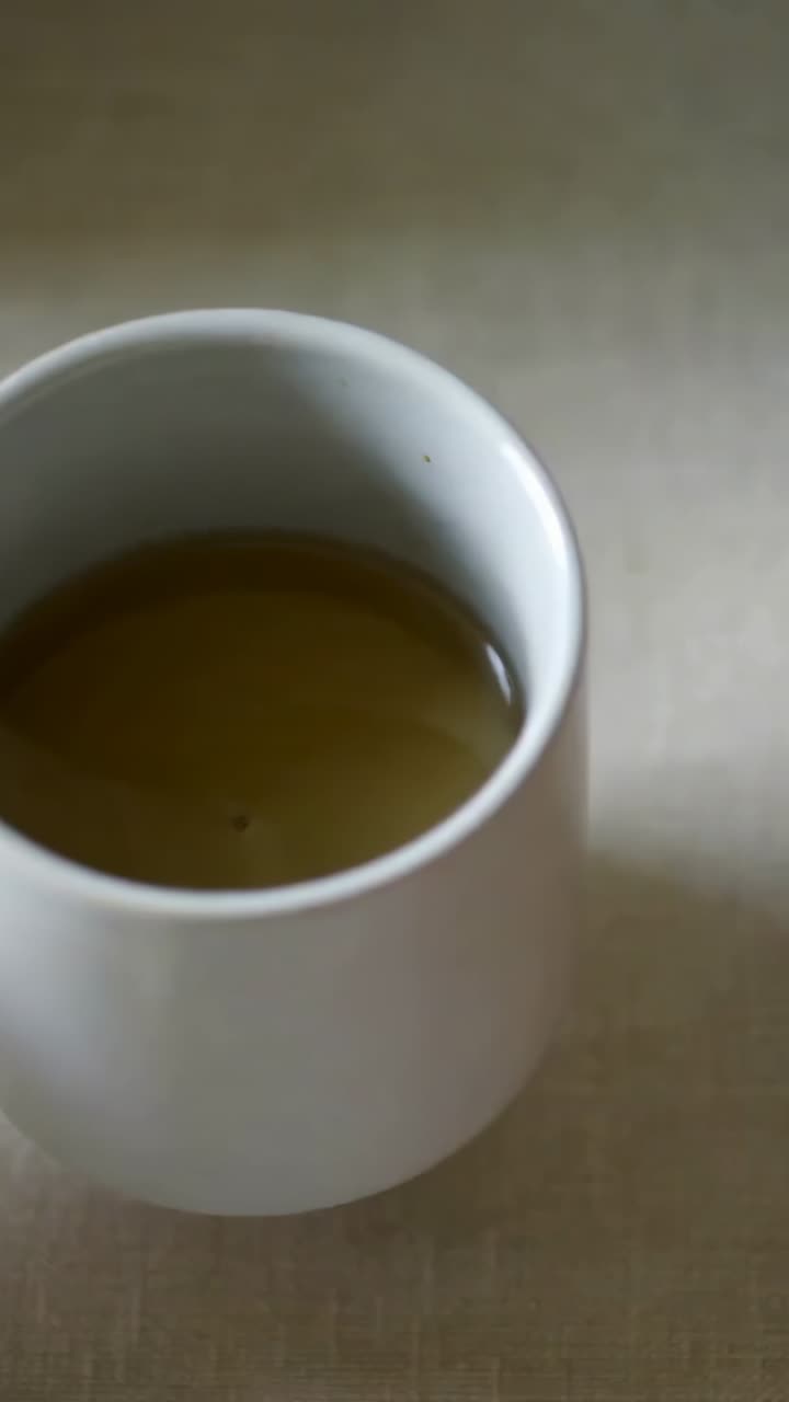 Vertical video: Holding mug, hand pinching loose tea leaves, pouring steel jug at table to brew tea