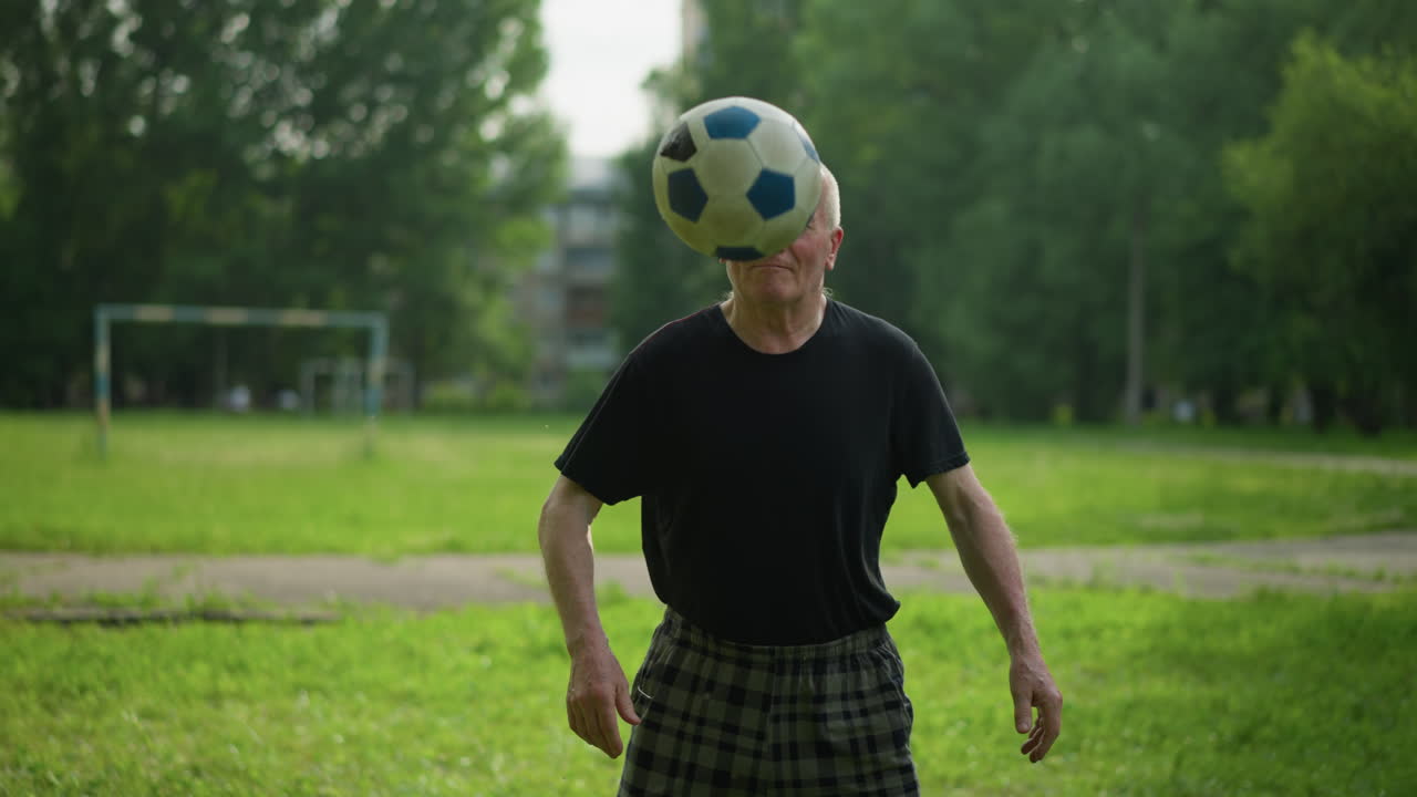 un anciano juega hábilmente con una pelota de fútbol en un campo de hierba exuberante, equilibrándola sin esfuerzo en su rodilla, con árboles y un poste de portería lejano en el fondo