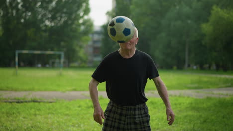 an elderly man skillfully plays with a soccer ball in a lush grassy field, effortlessly balancing it on his knee, with trees and a distant goalpost in the background