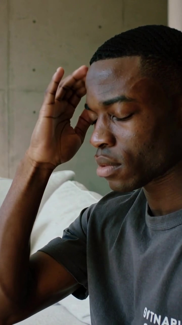 Vertical video: Pausing raising hand man in tee reflecting touching brow on sofa in living room