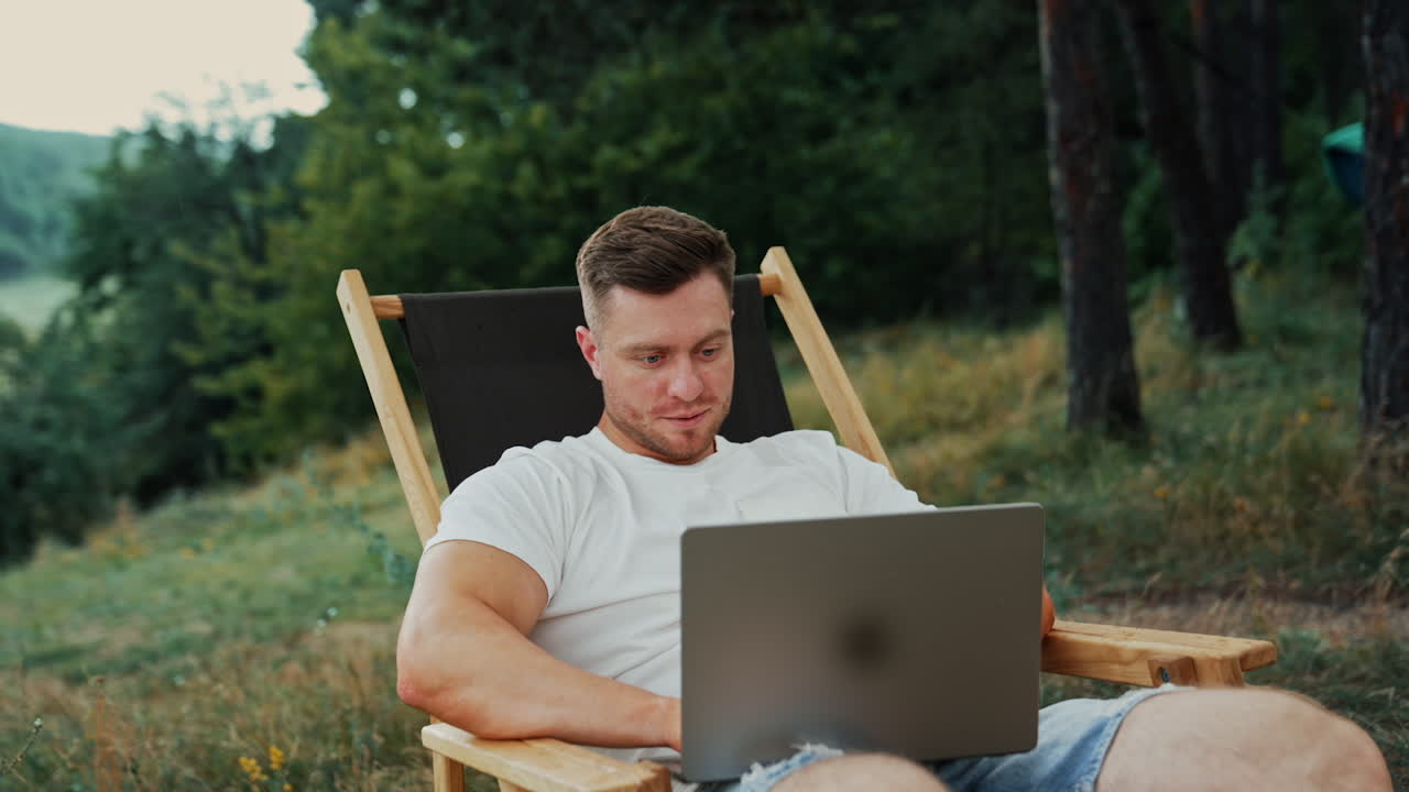 Mid-aged Caucasian man focused on his laptop. Freelancer works in the nature sitting in the folding chair.