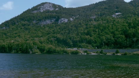 A-granite-mountain-covered-in-vegetation-stands-above-a-calm-lake-against-a-blue-sky-as-the-camera-pans