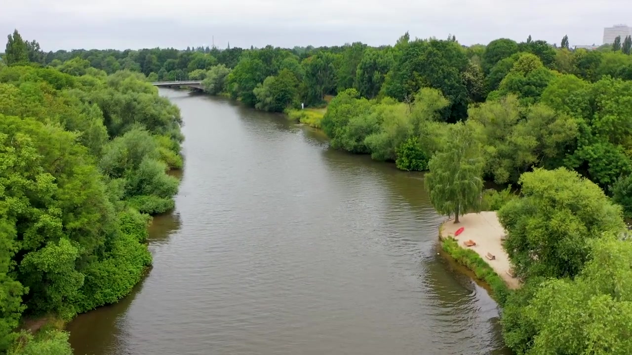 Cinematisk luftfoto af Leine-floden, der løber gennem frodigt grønne træer i Hannover, Tyskland, med en lille sandstrand.