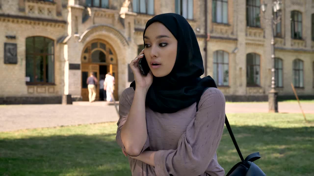 Young sweet muslim girl in hijab is talking on phone and hand up in daytime in summer, building on background, religiuos concept, communication concept