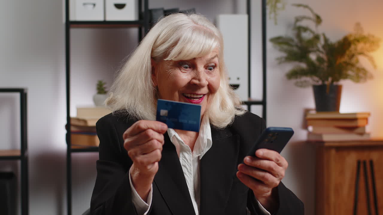 Happy caucasian senior woman using credit card and smartphone for online shopping payments at office