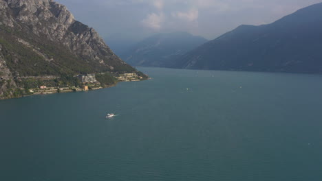 Toma-Aérea-Rápida-Del-Majestuoso-Paisaje-Del-Lago-Garda-Con-Un-Gran-Barco-De-Transporte-Y-Gente-Practicando-Deportes-Acuáticos