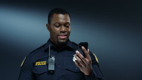 young cheerful handsome policeman in uniform and in walkie talkie smiling and having videochat on the smartphone via web cam on the dark wall background