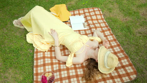 Woman-in-Yellow-Dress-Taking-a-Nap-Lying-on-Picnic-Blanket-with-Straw-Hat-Covering-Face-Relaxing-in-A-Park-Green-Lawn---Orbit-Top-View