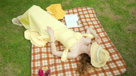 woman in yellow dress taking a nap lying on picnic blanket with straw hat covering face relaxing in a park green lawn - orbit top view