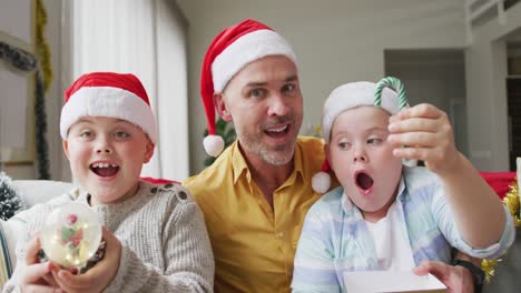 Portrait-of-caucasian-father-and-two-sons-holding-a-snow-globe-and-candy-cane-sitting-on-couch