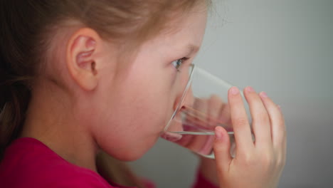 close up side view of cute young girl wearing pink shirt drinking water from clear glass indoors capturing simple healthy daily routine with focus and innocence in bright casual home environment