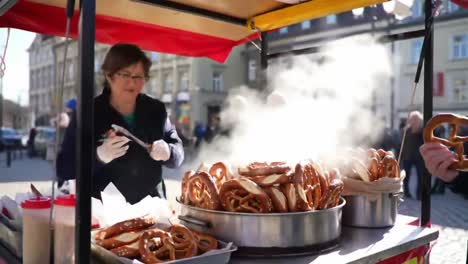 a busy street food scene featuring a vendor serving freshly made pretzels amidst a crowded market with steaming delicacies on display