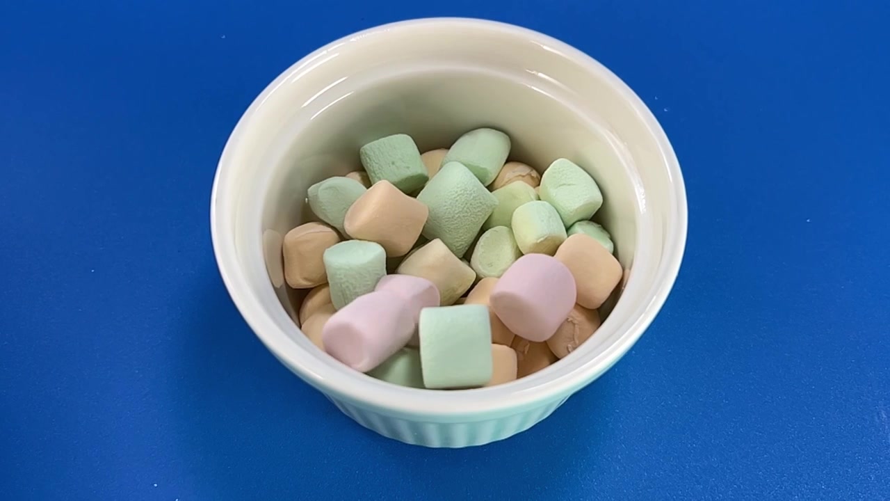 Marshmallow colorati cadono in una ciotola bianca su sfondo blu, creando un'azione alimentare giocosa con colori pastello tenui e forme variegate