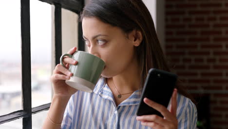 mujer bebiendo café y usando el teléfono inteligente junto a la ventana
