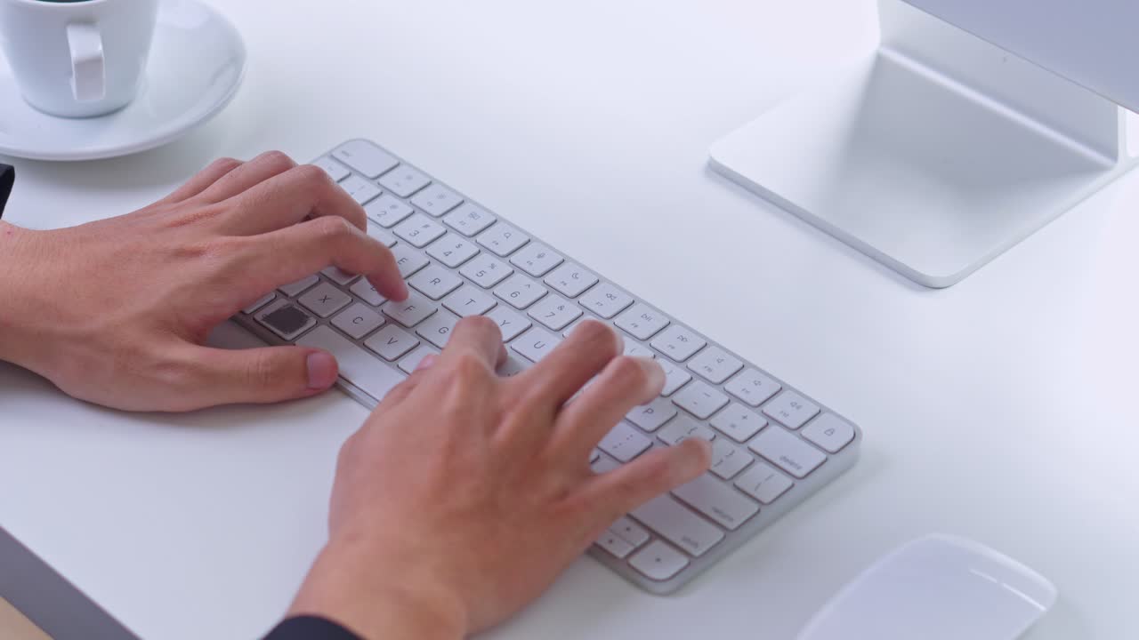 Close-up of male hands typing quickly on modern wireless keyboard at white office desk with coffee