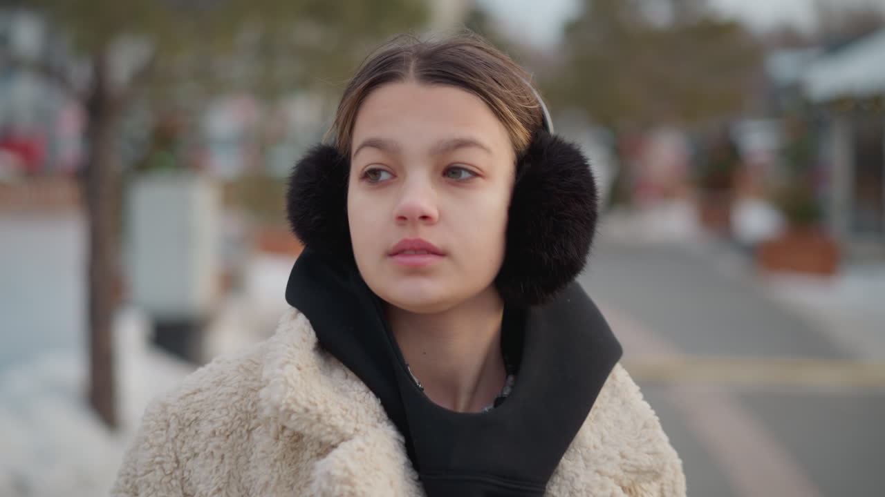 Young girl in warm beige winter jacket with black hoodie and fluffy earmuffs turns around calmly on snowy street, blurred background with decorated trees, snow-covered ground