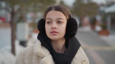 young girl in warm beige winter jacket with black hoodie and fluffy earmuffs turns around calmly on snowy street, blurred background with decorated trees, snow-covered ground