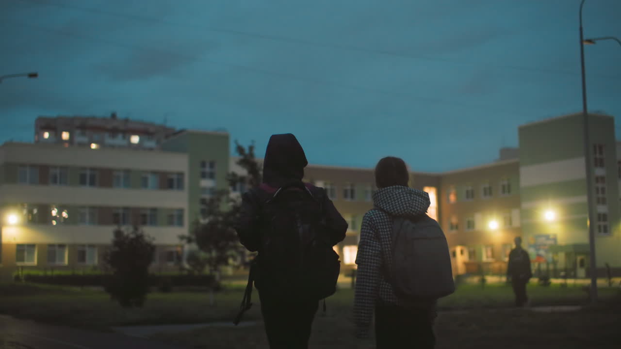 Two children with backpacks walking toward school building in early morning light, silhouettes visible against blue sky, exterior lights illuminating scene, urban neighborhood quietly beginning daily routine