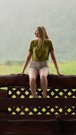 elegant woman in green t-shirt looks aside sitting on terrace log fence against large lush meadow and mountain silhouette on rainy day slow motion