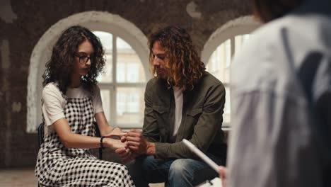 A-guy-with-curly-hair-in-a-green-jacket-and-a-brunette-girl-with-curly-hair-in-glasses-hold-hands-and-communicate-with-a-professional-psychologist,-a-girl-in-a-shirt,-in-a-brick-building