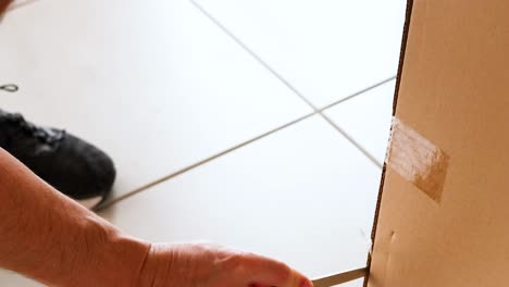 close-up of hands using a knife to cut taped edges of a cardboard box on a tiled floor.