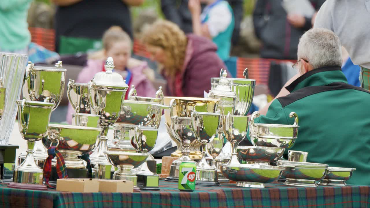 Judges assess numerous trophies at outdoor Highland Games, with people and teamwork visible