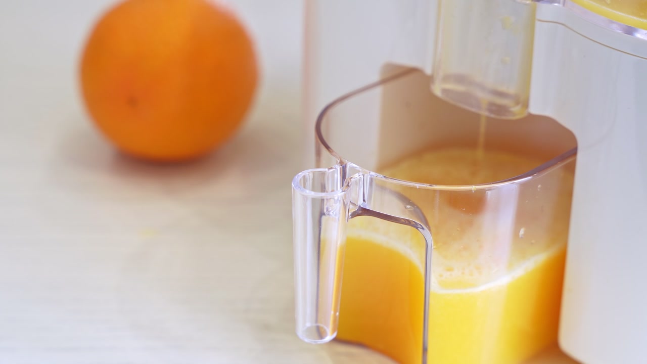 En hvid elektrisk juicer udvinder frisk økologisk appelsinjuice i en klar kande. Filmisk nærbillede i et lyst køkken med lav dybdeskarphed til sundheds- og wellnessreklamer.