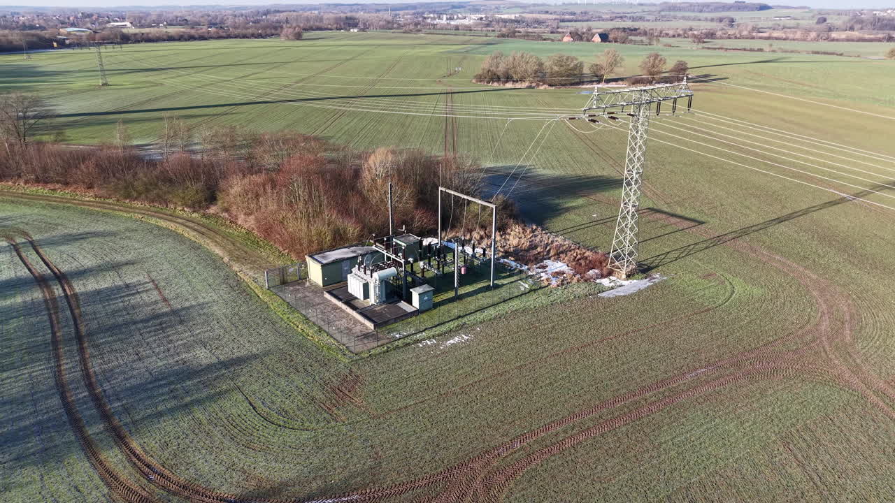 Vista aerea da drone di una sottostazione elettrica rurale collegata a un traliccio di trasmissione ad alta tensione, circondata da campi agricoli e vegetazione invernale. La scena evidenzia la rete elettrica fondamentale