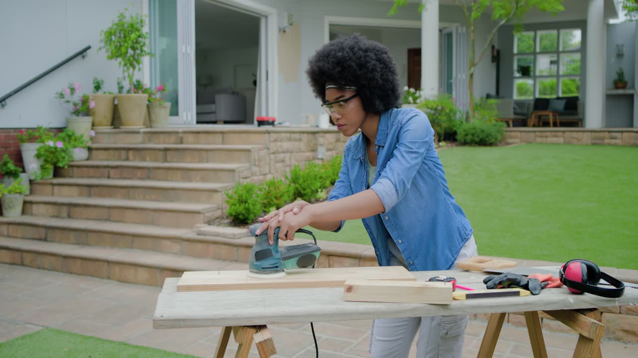 Femme afro-américaine utilisant une ponceuse, ponçant une planche sur un banc de terrasse, lissant le bois pour un projet.