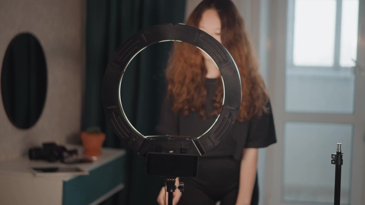 Woman with long hair stands behind illuminated ring light while tightening smartphone mount on tripod, hands positioned on mount and slight forward lean indicating active setup and focused