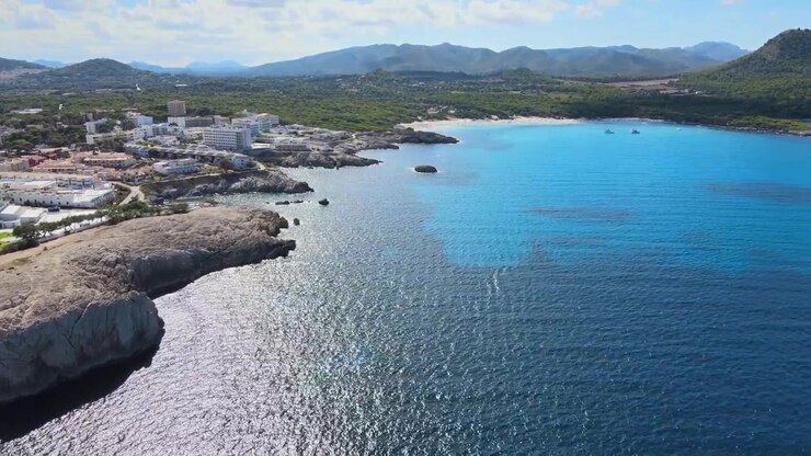 Experimente as vistas deslumbrantes de Cala Agulla em Cala Rajada, Maiorca, onde as águas azuis claras encontram a costa rochosa e a paisagem exuberante e vibrante sob o sol.