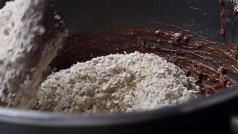 Close-up-on-wooden-spoon-,-stirring-liquid-chocolate,-nuts,-cereal-and-oats-in-black-pot,-making-homemade-energy-bars-120fps