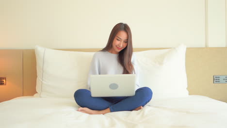 smiling asian woman sitting on bed with laptop