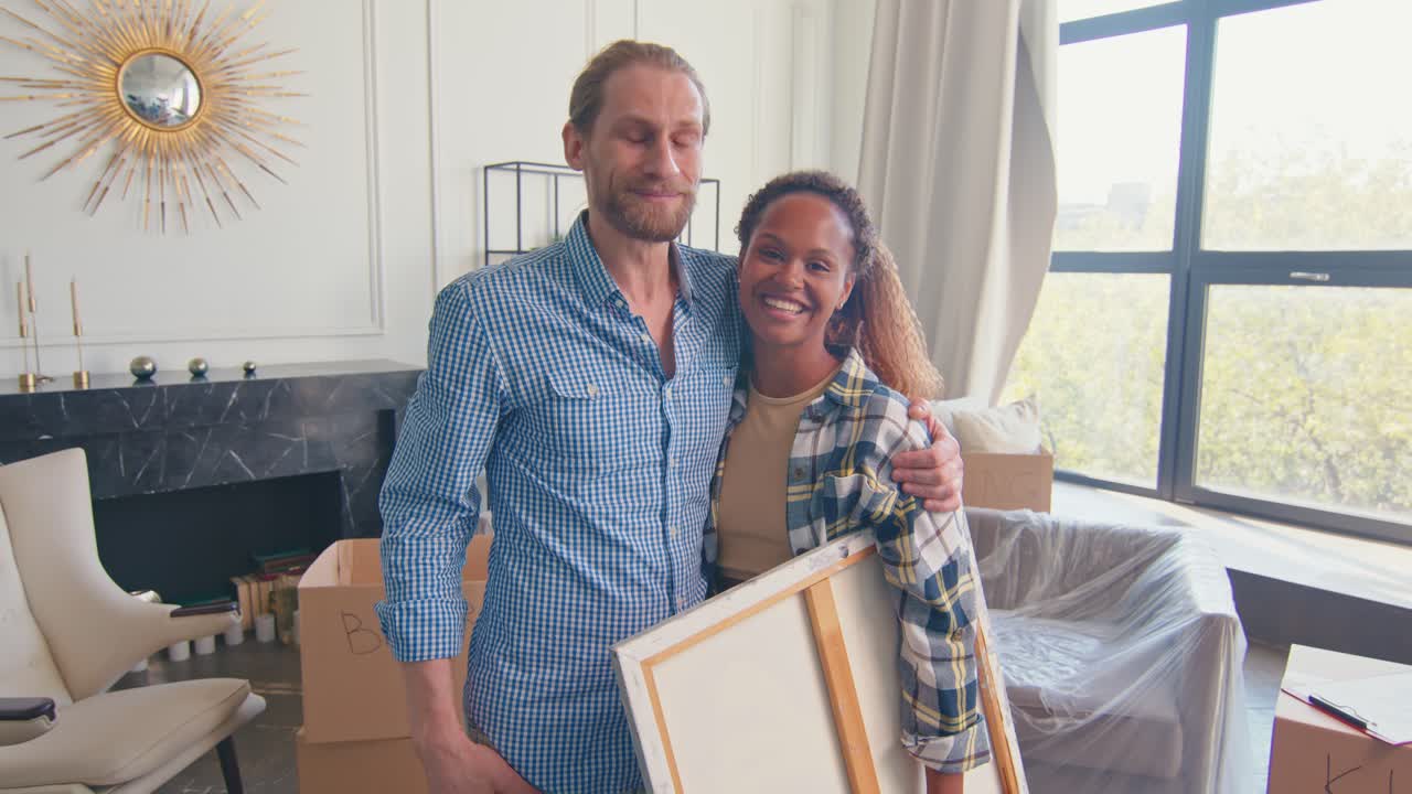 una pareja joven y diversa abrazándose después de mudarse y sosteniendo cuadros en casa.