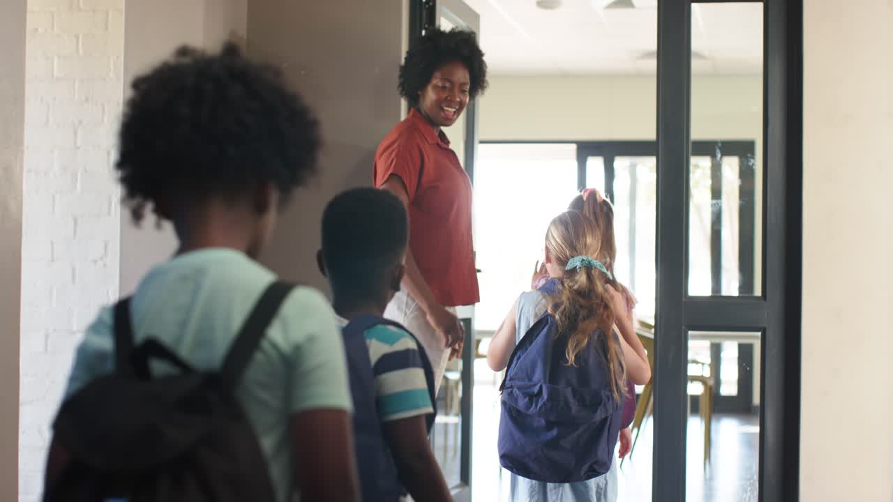 Diverse children arriving African American woman greeting guiding backpacks via door checking list