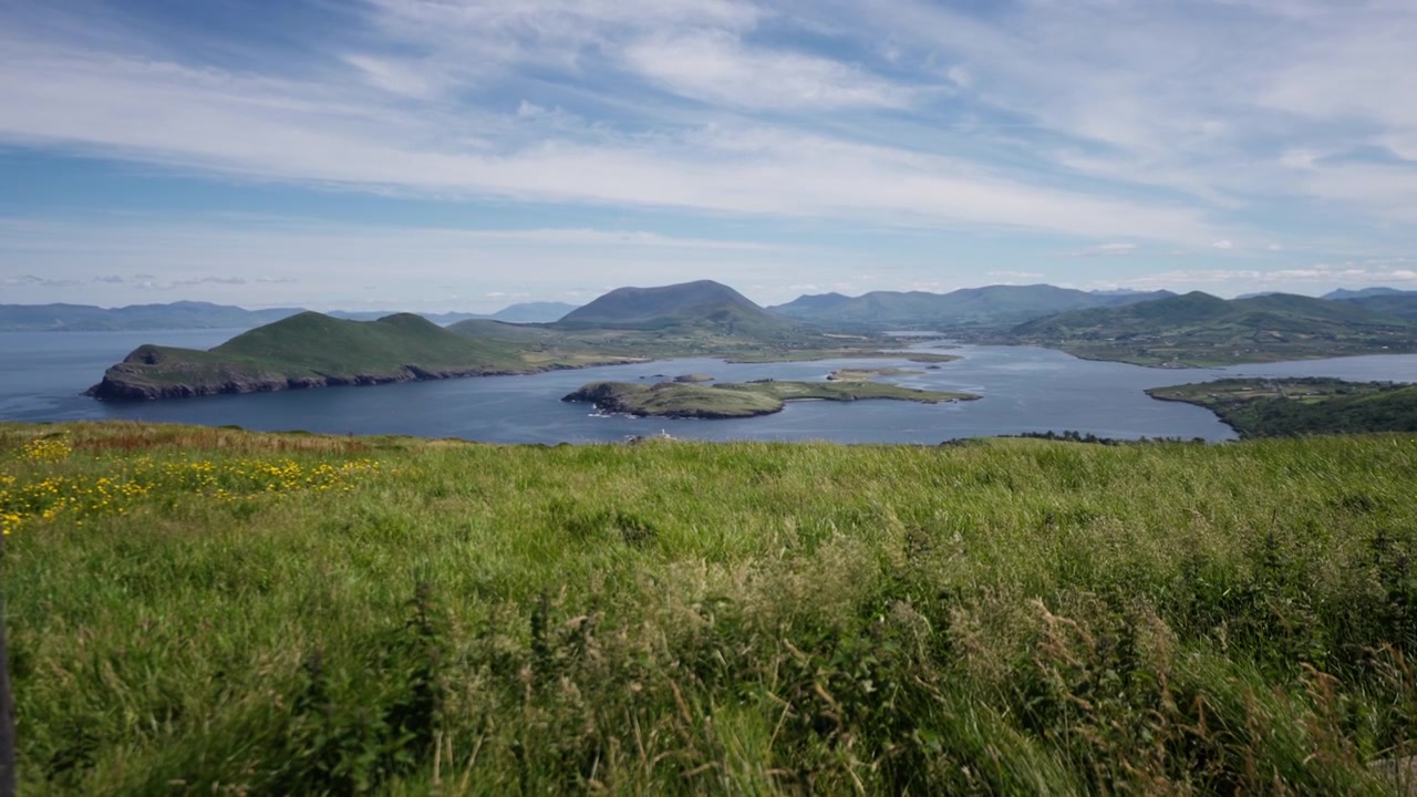 Vid utsikt over bølgende grønne åser og kystnære øyer under en blå himmel med spredte skyer og fjerne fjellkjeder.