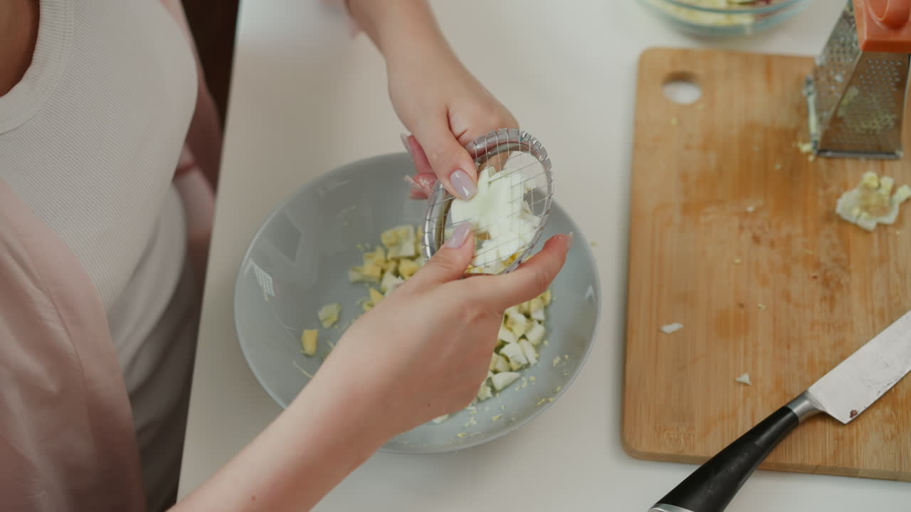 Student using small net egg smasher to crush boiled egg into bowl, working carefully with both hands in bright kitchen, surrounded by kitchen tools, knife, grater, and other fresh ingredients