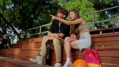 two teenagers sitting on a bench at a basketball court, talking and laughing